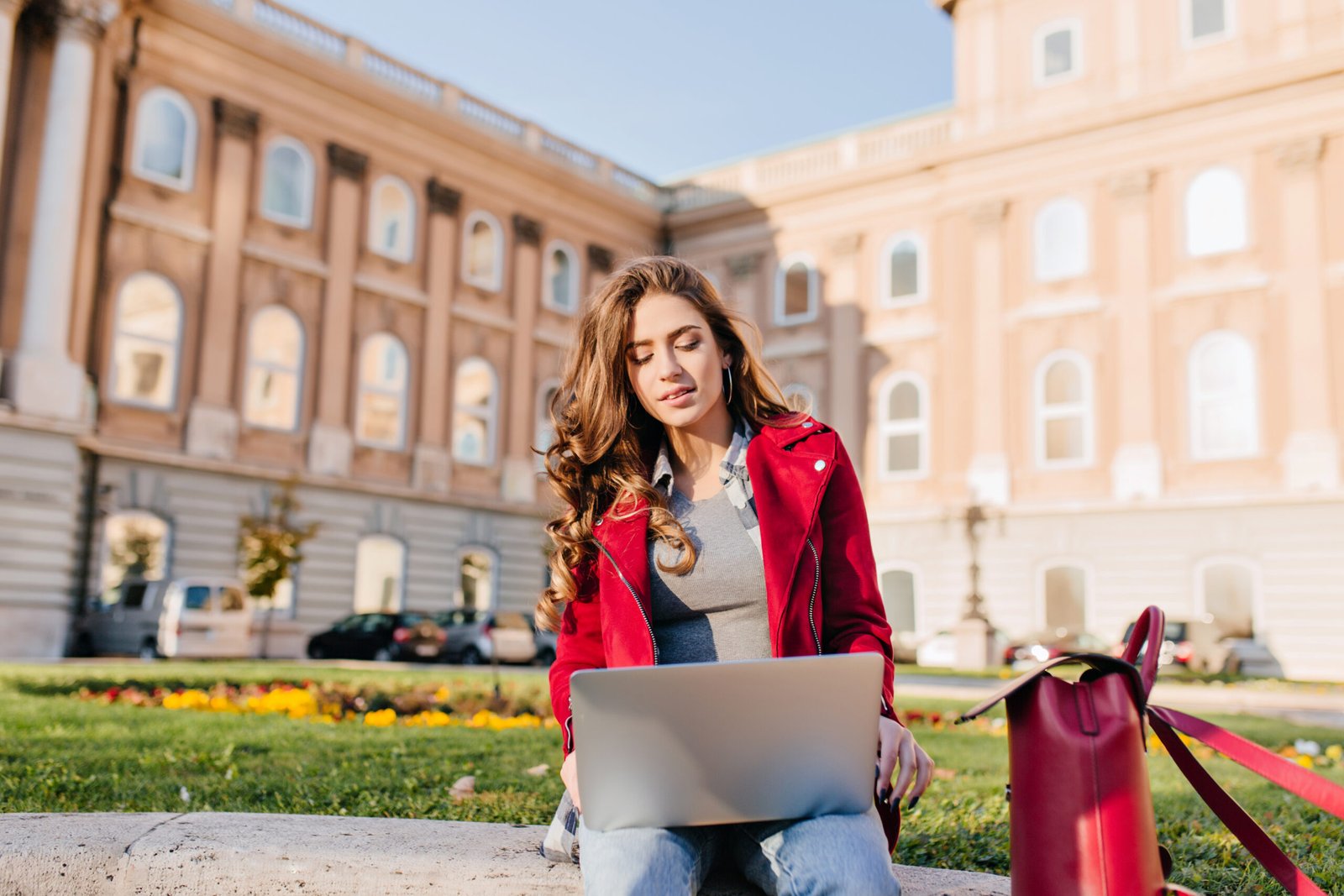 Outdoor portrait of serious curly female student sitting with laptop on the ground. Busy brunette girl in red jacket working with computer in front of college in warm day.